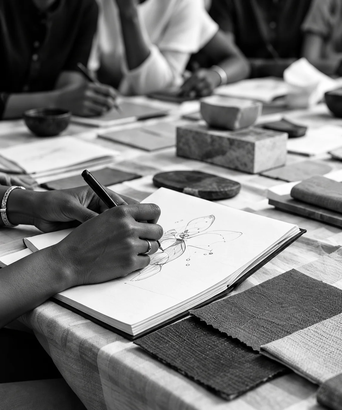 Hands sketching in a notebook at a table with fabric samples while others work around it